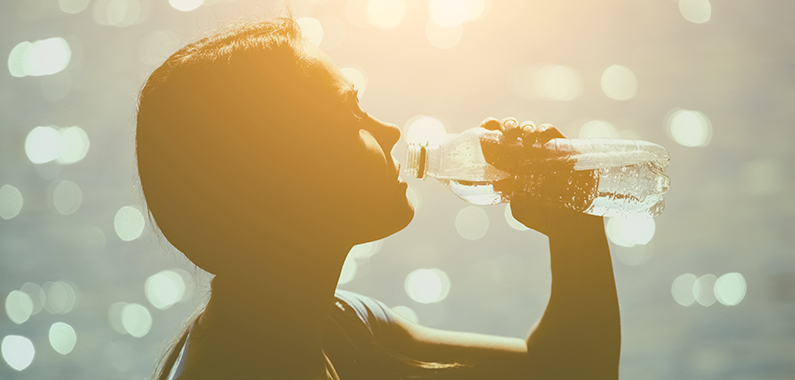 Silhouette of a young female athlete in tracksuit drinking water from a bottle on the beach in summer during morning exercises. Sport and healthy lifestyle.