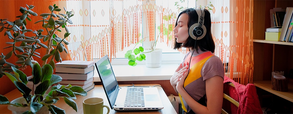 Woman with her hands on her chest meditating with headphones on in an office in front of the computer