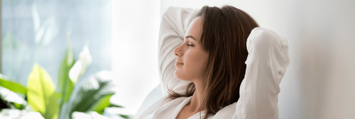 Woman relaxing with her arms behind her head and eyes closed, sitting in a sunny room.