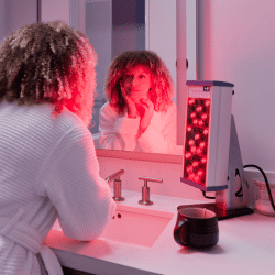 Woman using the Clearlight PERSONAL Red Light Therapy Tower while admiring the red light on her skin in a bathroom mirror