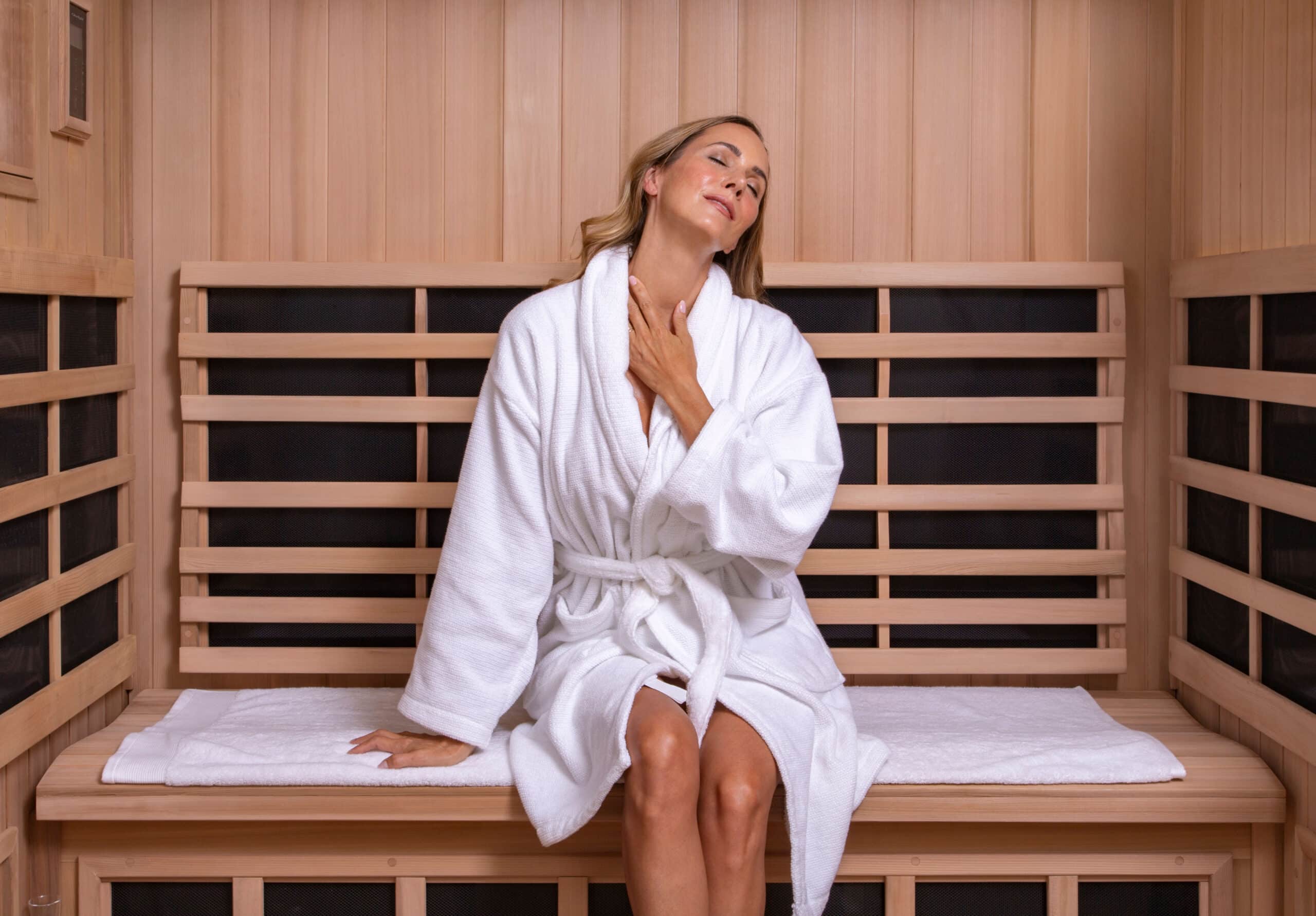 Woman in a white bathrobe sits in a wooden sauna, eyes closed, hand resting on her chest in a relaxed pose.