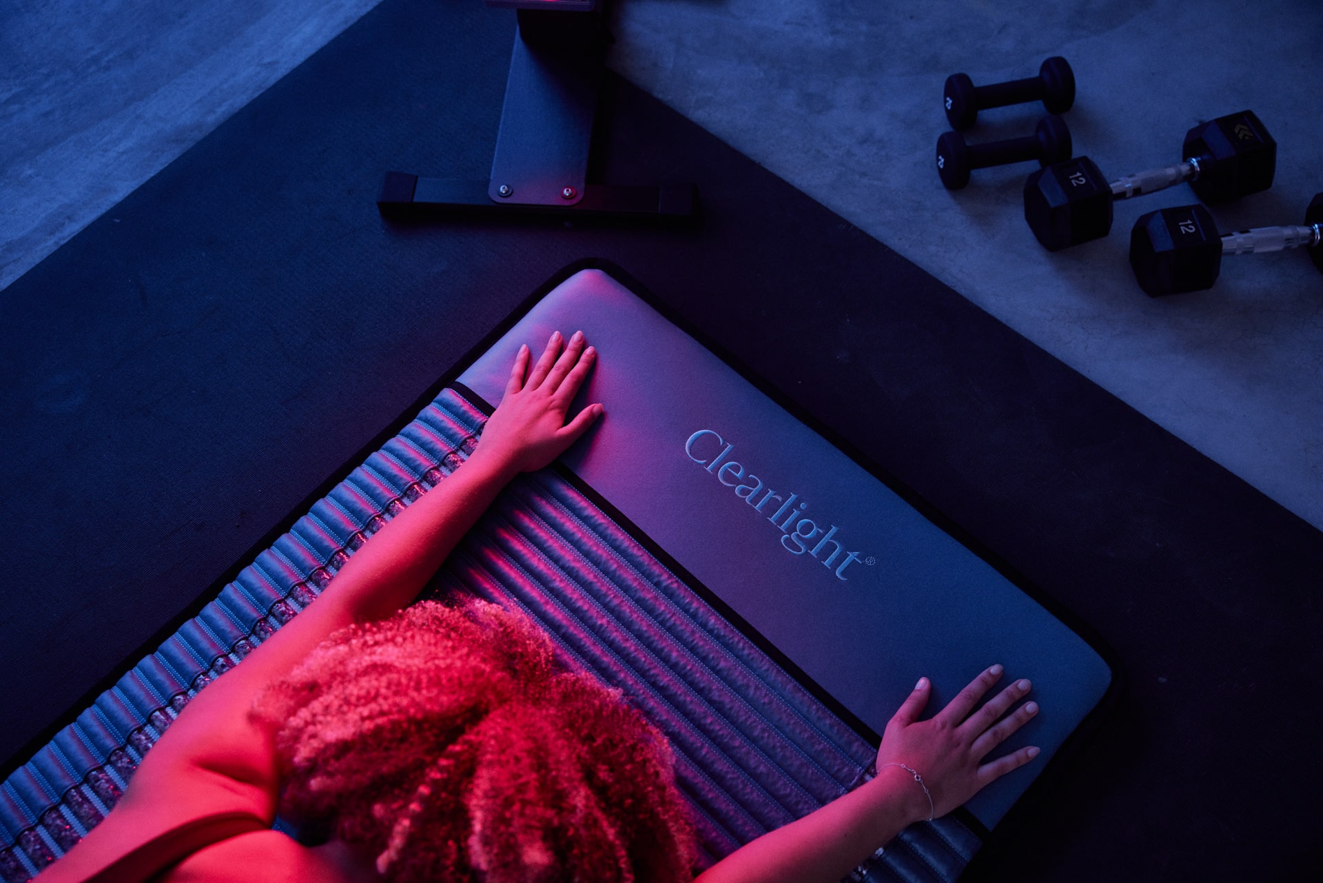 Overhead view of a person with curly hair on a ClearLight exercise mat, hands pressing on the mat while blue-pink gym lighting shines on weights nearby.