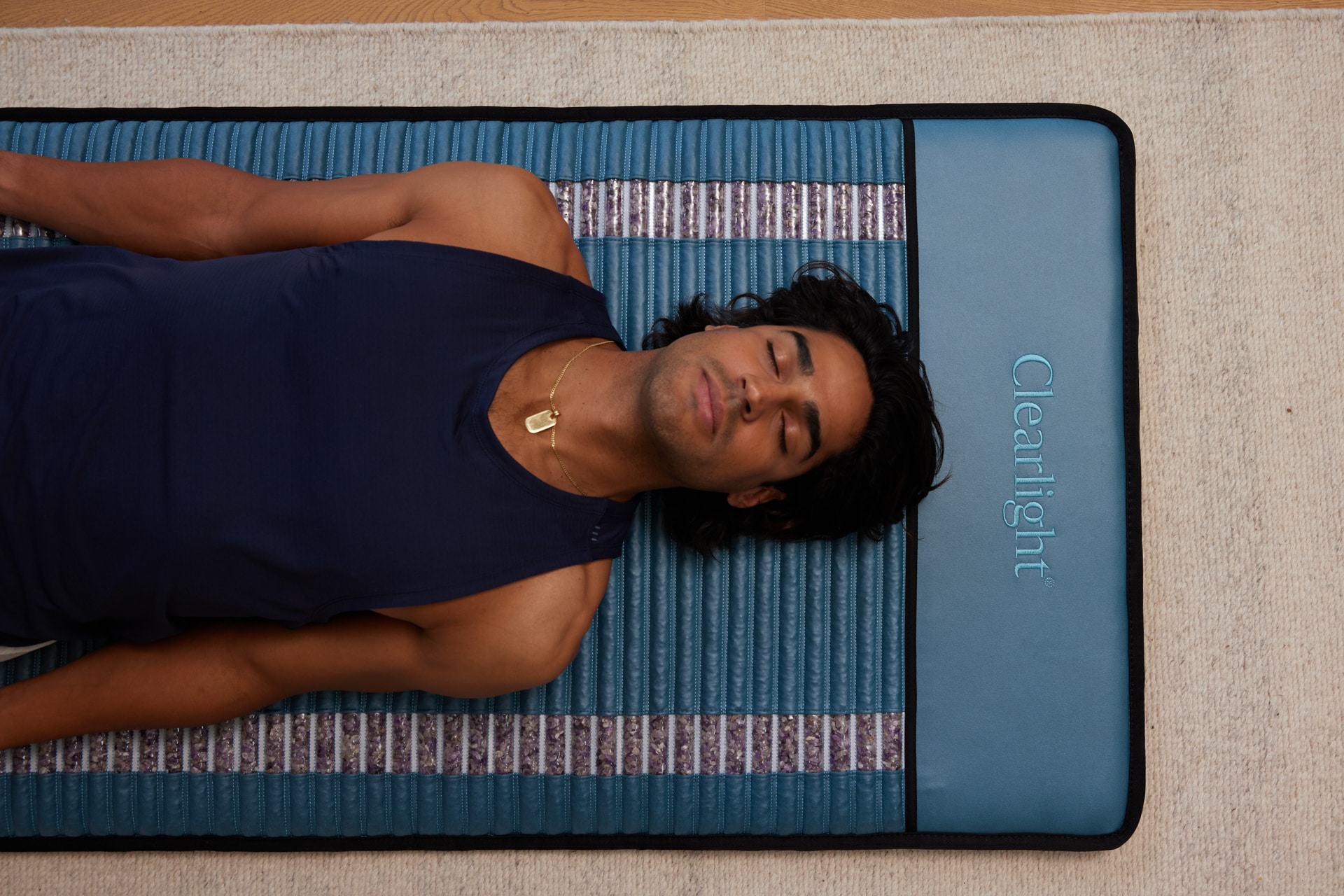 Man wearing a navy sleeveless top lies on his back, eyes closed, on a blue padded exercise mat on a beige carpet.